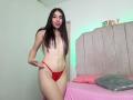 SophiaStorm, topless, with long brown hair, small tits, red thong, standing in a bedroom, smiling, hand on hip, pink bed with gray pillow, headboard with "TWO!" sign.