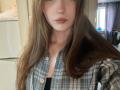 EleanoraMirabal with long brown hair, wearing a plaid shirt, buttoned, showing her chest, pinkish lips, slight smile, and a thin necklace in a softly lit room.