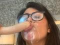 CatalinaReiyna with black glasses, dark hair, and tan skin, sucking a large, circumcised, erect dildo. Her face and mouth are covered in thick, white cum.