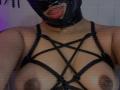 LoreDuval wearing a black mask with red lips, strappy black harness revealing her tits, with visible nipples and dark areolas, against a blurred background.