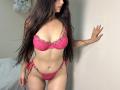 Tisia with long dark hair, wearing pink lace bra and matching thong, standing against a wall, right hand on it, showing her tits and pussy, with a slim, curvy body.