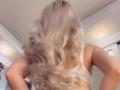 MademoiselleBella's back, showing long, wavy blonde hair, white bra, and her ass, with her arms slightly raised, in a softly lit room.