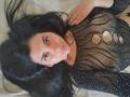 JeneKurz with long black hair, wearing a black, see-through, mesh top with cut-out, showing small, round tits. She's lying on a beige surface, looking at the camera.