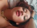 VioletaSwitch has curly red hair, green eyeshadow, and her tongue out, lying on a bed with her hand in her hair, showing her pink lips and green bracelet.