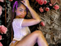 SkinnySamantha, avec des cheveux tressés, des rubans violets, portant un haut blanc transparent, assise les jambes levées, montrant sa vulve, entourée de roses roses et de journaux..