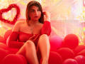SaraWestt with red, sparkly off-shoulder dress, sitting in pink balloons, wearing a tiara, showing small tits, with one hand in her hair, looking at the camera.