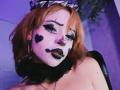ViolettClay has orange hair, white face paint with black heart and lip marks, dramatic eyeliner, black lipstick, and a black ruffled choker, sitting in a purple-lit room.