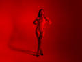 LauraJackson is standing nude, her hands on her hips, with long dark hair, black high heels, and a red light illuminating her. She has small tits, a tattoo on her right thigh.
