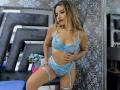 CloeRouss in blauwe kanten lingerie, met een korset en kousen, die haar borsten en vulva laat zien, met blond haar, tatoeages en rode lippenstift, staand in een slaapkamer..