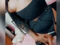 DenyChantal's black strapless bodysuit with metal buckle, showing her tits, has her hand with tattoos on her thigh, long black hair, pink bed, white stuffed toy.