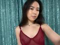 MiiaVixen with medium brown skin, long black hair, wearing a sheer red bralette, showing small tits, subtle makeup, and a black necklace, standing in front of teal curtains.