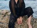 Niferttiti, wearing a black lace dress, sits on a rock with her legs apart, showing red-painted nails and a bare foot, her long brown hair covering her face.