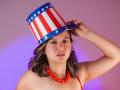 ArianaBlonde wears a red, white, and blue striped top hat with stars, a red bead necklace, and a red strapless top, holding the hat with a slightly open mouth.