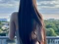 Tisia's long, straight brown hair covers her back, wearing a gray tank top, standing on a balcony with her ass facing the camera, cityscape in the background.