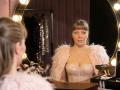 VeronikaGoldy with her tits in a pink, sparkly, feathered dress, staring into a mirror, holding makeup brushes, in a warmly lit makeup room.