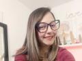 ArianaBlonde with glasses, smiling, showing her red lipstick and slightly visible teeth, wearing a red top, has shoulder-length brown hair, in a well-lit room.