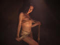 TriciaBarnes with bare tits, bound in rope, wearing a shiny gold skirt, standing in dim light, holding rope, tattoo on left side, dark background, sexy, confident pose.