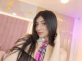 NaiaSanders with long black hair, pink plaid tie, nose ring, and fishnet top, smiling at the camera, hand in hair, pink and white background.