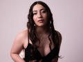 AnnFisher has wavy dark brown hair, a cute face, and small tits in a black top. She's wearing a necklace with a small pendant. Soft light, neutral background.
