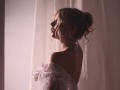 RosaVance with blonde hair in a bun, wearing a sheer white dress with flower accents, facing left, showing her bare back and neck, soft light, elegant, sexy.