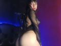 Boorbuja with black fishnet top, exposing tits and ass, standing with back to camera, looking over shoulder, in dim blue-purple light, with long black hair and bangs.