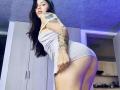 SophiaMarx in a tight white dress, pulling it up to expose her ass, tattoos on her arm and side, long black hair, looking over her shoulder. Bedroom setting.