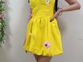 MiraMixi in a bright yellow dress with pink and white flower embroidery, black straps, and red nails, standing with her hand on her hip.