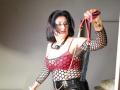KarlaHartley with black fishnet top, red lace bra, black leather pants, holding red and black bondage straps, wearing black ball gag, looking annoyed, medium-length black hair, light skin.