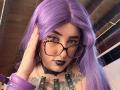 LilithDark69 with purple hair, black glasses, goth makeup, black lipstick, tattoos, leather choker, purple wrist cuffs, hand in hair, wooden ceiling in background, looking at camera.