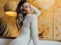 MikyWayxy in a white, long-sleeve, knitted dress, showing her ass, looking back with a hand in her wavy blonde hair, under two hanging woven lamps.