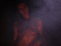 TriciaBarnes with tied-up boobs, foggy face, and rope across her tits, standing in dark, smoky red light. She has a serious expression, looking directly at the camera.
