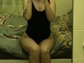 Olieri is sitting on a bed wearing a black one-piece, her hands covering her tits, showing a tattoo on her left arm, her pussy is covered, her ass is visible.