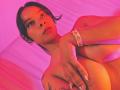 Yurimy with tan skin, black hair, and tattoos, holding her large tits with one hand, wearing a jeweled bracelet, under pink and red lighting.