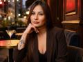 AnaEoxo has long brown hair, a tattoo on her right wrist, and is wearing a brown blazer over black lace. She's sitting with a martini glass in the background.