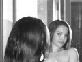 Fotografia in bianco e nero di MicheleJinkins con i capelli lunghi, che guarda il suo riflesso in uno specchio, mostrando le spalle scoperte e un sorriso appena accennato. Il seno è visibile nel riflesso..