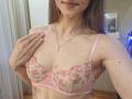 MilaYanis with pink floral bra, showing tits, long brown hair, pink nails, gold necklace, hand on chest, small tattoos, wooden floor, smiling, indoor, daytime, casual, sexy.