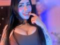 Kkole with long black hair, smiling, finger in mouth, brown ribbed tank top, big tits, tattoos on arm, blue and pink lighting, nose ring, makeup, and long eyelashes.
