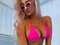 MademoiselleBella with big, pink bikini top showing her large, perky tits and wearing pink sunglasses, has long, wavy blonde hair, and a cross necklace, looking sexy.