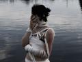 JanetWiedemann has black hair, wearing white bandage-like wraps over her tits and arms, covering her mouth, standing by a reflective water surface. She's wearing a pearl necklace.