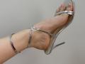 Tattiya's foot in a shiny silver high heel, showing her toes and ankle, with the heel strap wrapped around her ankle. Her foot is slightly raised.