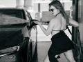 Milagres with tits exposed, wearing sunglasses, black skirt, and halter top, washing a black car with a hose at a gas station, showing her ass. Black-and-white photo.