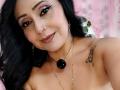 Zaria69 with dark hair, makeup, nose ring, earrings, necklace, and tattoo on right shoulder, smiling with pink lipstick, showing bare shoulders. Boobs not visible.
