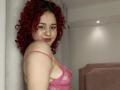 RosalieVittoria with curly red hair, pink lace bra, showing her tits, smiling at the camera, standing against a white wall, soft pink headboard in the background.