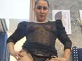 SilverCool69, medium-skinned woman with dark hair in a bun, wearing a sheer black top revealing small tits and a black lace thong, hands on knees, looking at camera.