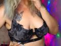 LilithGreenn with large tits in a black lace bra, showing off tattoos on her chest. She's holding her phone, with blonde hair and blue nails.