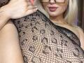 BaeonLive wearing black fishnet stockings with floral pattern, showing her tits, holding her leg up, blonde hair, glasses, and pink lipstick. Her hand has a small tattoo.
