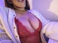 AntoniaBrunni with large tits in a red lace bra, open white cardigan, and her tongue out. Her chest is prominently displayed, showing her cleavage and bra.