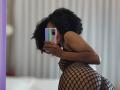 SnackTime with curly black hair, wearing a black fishnet bodysuit, bent over, showing her ass, taking a selfie with a colorful phone in a bedroom.