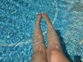 Barbarica's legs are submerged in a blue-tiled swimming pool, water rippling over her skin, toes pointing upward, sunlight creating wavy reflections on her pale skin.