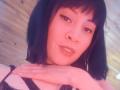IssisDiosa with black hair, subtle makeup, and a relaxed smile, resting her chin on her hand, wearing a black lace top, showing her tits. Soft pink light.